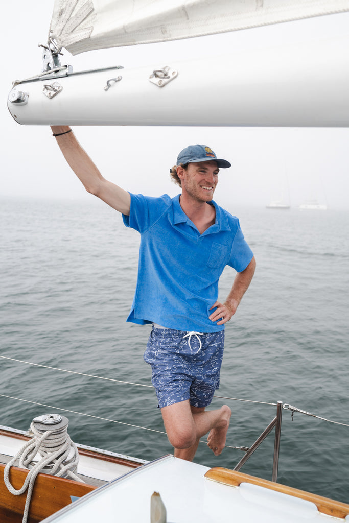 Man on a sailboat in a blue Terry polo shirt and blue printed swim trunks.