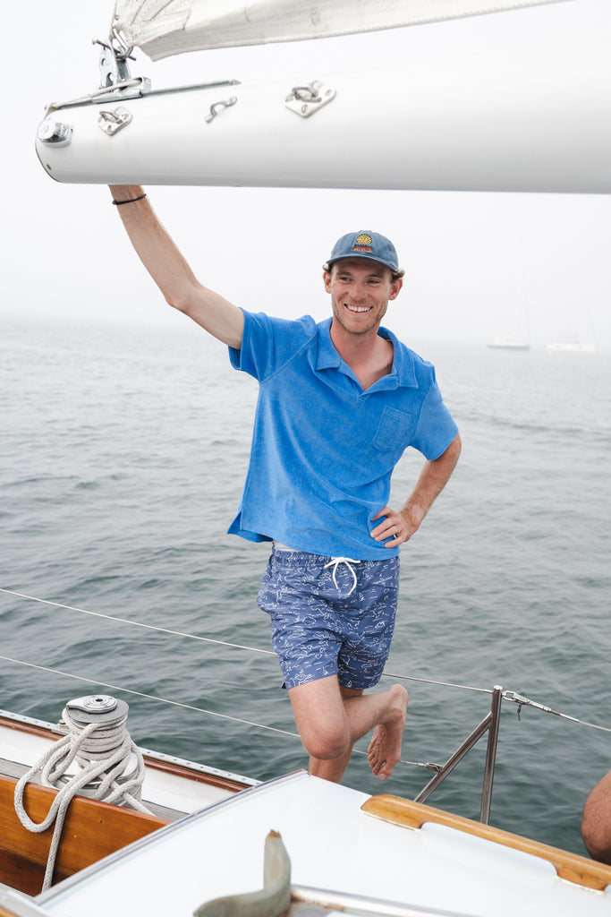 Man on a sailboat in a blue Terry polo shirt and blue printed swim trunks.