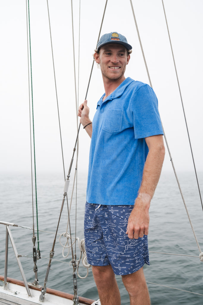 Man on a sailboat in a blue Terry polo shirt and blue printed swim trunks.