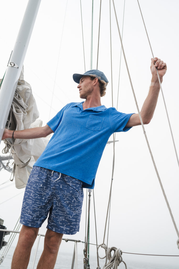 Man on a sailboat in a blue Terry polo shirt and blue printed swim trunks.
